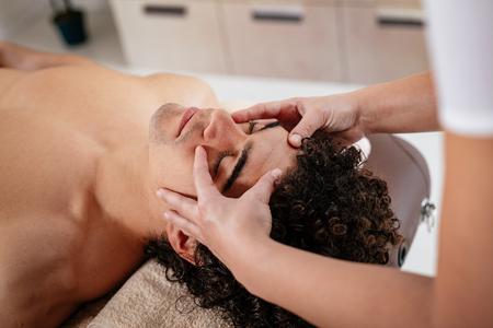 Close-up of a handsome healthy young man enjoying relaxing facial massage at beauty salon.の写真素材
