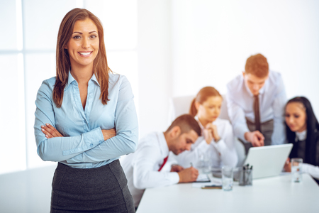 Successful young businesswoman standing proudly in the office and looking at the camera. Her young colleagues working at laptop on desk.の写真素材