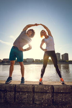 Young happy couple training outdoors by the river, stretching together on the wall at sunset.の写真素材