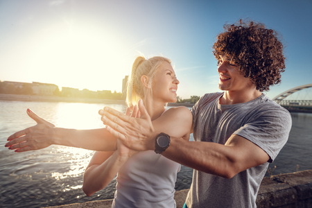 Young fitness couple doing workout by the river in a sunset. The woman is stretching her shoulder and the man support her.の写真素材