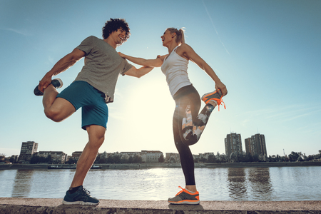 Young happy runners training outdoors by the river, stretching legs at sunset.の写真素材