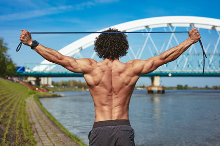 Rear view of a bodybuilder with naked torso, who is doing strong fit body training with elastic rubber band, near the river.の写真素材