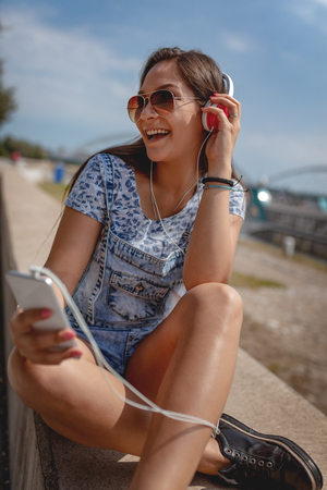 A beautiful cheerful young woman listening music from your phone and enjoying on the city rivershore on a beautiful summer day.の写真素材
