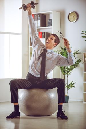 The young businessman sitting in the office on pilates ball and doing exercise with dumbbells.の写真素材