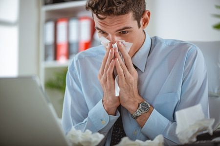 Young businessman blows his nose while working at his laptop in the office.の写真素材