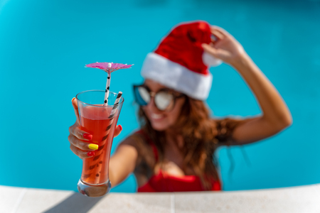 Young beautiful woman near the swimming pool in Santa Claus hat celebrating New Year and Christmas in hot country with glass of cocktail.の写真素材