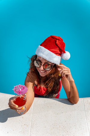 Young beautiful woman in the swimming pool in Santa Claus hat celebrating New Year and Christmas in hot country with glass of cocktail.の写真素材
