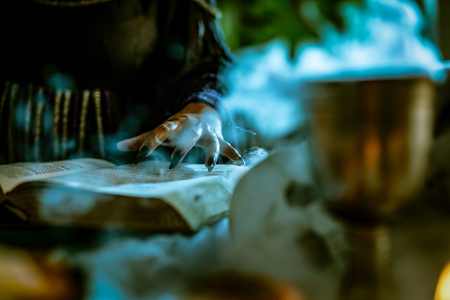 Close-up of a witch's hand with pointed black nails on smoky book of magic recipe and with a magic wand.の写真素材