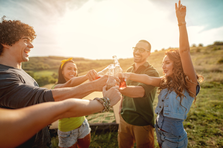 Happy young friends enjoy a sunny day in nature. They're clinking beer bottles, laughing and talking happy to be together.の写真素材