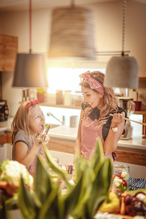 Beautiful woman with her daughter looks happy while preparing meals and tasting vegetable in the kitchenの写真素材