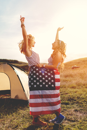 Happy young lesbian girl enjoys a sunny day in nature. They're wrapped in american flag in front a campsite tent and dancing.の写真素材