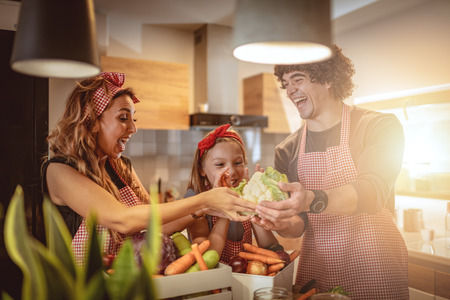 Cute little girl and her beautiful parents are cutting vegetables and smiling while making pickle in kitchen at homeの写真素材