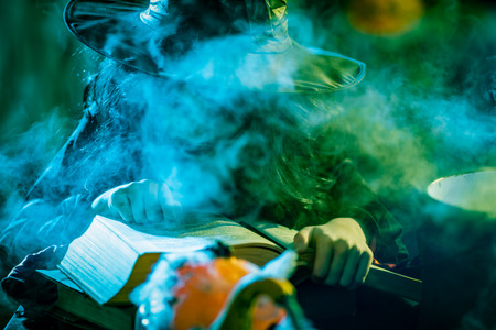 Witch with awfully face reading recipes of magic drink in creepy surroundings and smoky background.