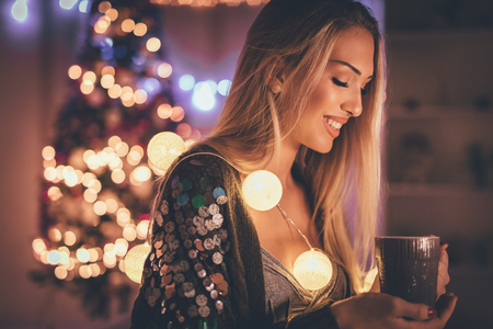 Cute young smiling woman holding cup of coffee, surrounded with Christmas bubble lights.の写真素材