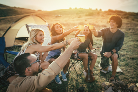 Happy young friends enjoy a sunny day at the mountain. They're laughing and grilling -roasting sausages on sticks over a campfire near tent.の写真素材