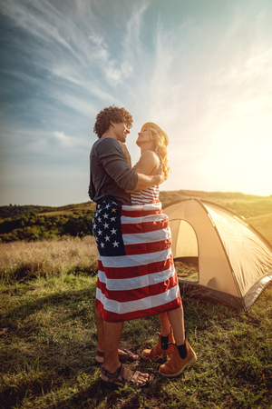 Happy young couple enjoys a sunny day in nature. They're hugging each other, wrapped in american flag in front a campsite tent.の写真素材