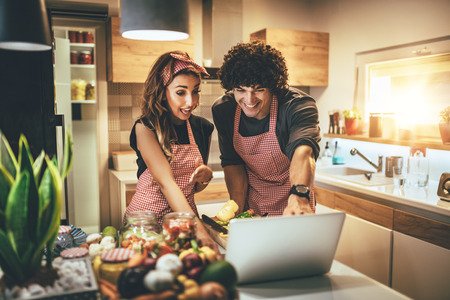 Young happy couple is enjoying and preparing healthy meal in their kitchen and reading recipes on...