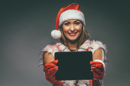 Beautiful young smiling woman in Santa Claus costume showing digital tablet and looking at camera. Selective focus.の写真素材