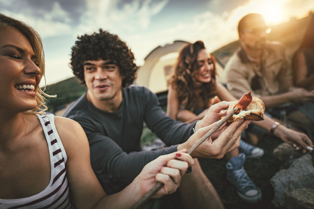 Happy young friends enjoy a nice sunny day in nature. They're grilling sausages, eating, laughing...