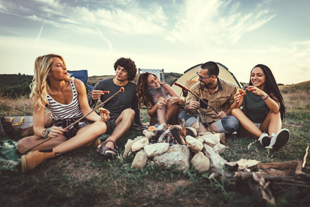 Happy young friends enjoy a nice day in nature. They're grilling sausages, eating, laughing and talking happy to be together.の写真素材