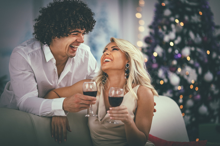 Young couple celebrating Christmas at home. They are relaxing with glass of red vine, covered with blanket on a sofa in an apartment.の写真素材