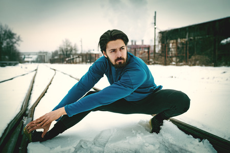 Active young man crouching and doing exercises in the public place among old railroad during the winter training outside in. Copy space.の写真素材