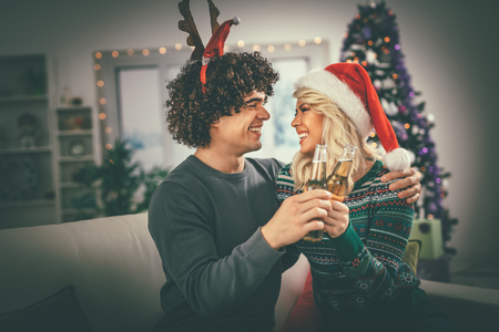 Young romantic couple hugging and celebrating Christmas at home, they are having fun and toasting with beer. Selective focus.の写真素材