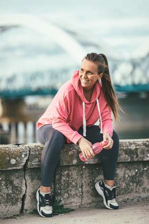 Young fitness woman is resting after hard training on the wall by the river bridge with bottle of the water.の写真素材