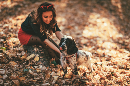 Cute young woman enjoying in city park in autumn colors. She is sitting on the ground covered with leaves and having fun with her dog.の写真素材