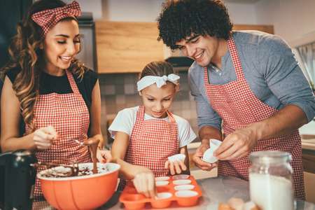 Happy parents and their daughter are preparing cookies together in the kitchen. Little girl helps to her parents to put paper cups into molds.の写真素材