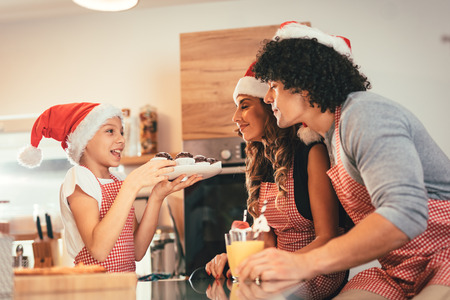 Happy parents and their daughter with santa's cap are preparing meal together in the kitchen. Little girl is holding plate with muffin offering to her mother and father.の写真素材