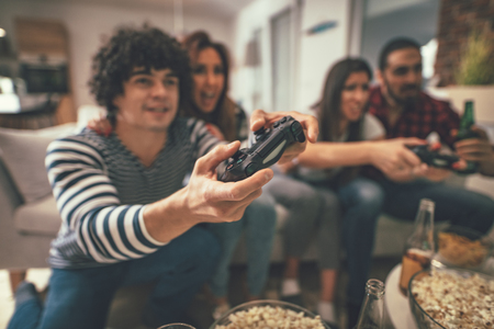 Excited friends are playing video games at home sitting on sofa with beer. They have great weekend...