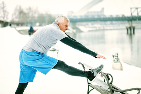 Active senior man athlete stretching leg on the bench and doing exercises by the river during the...