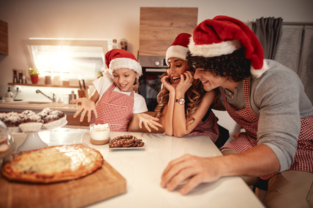 Happy parents and their daughter with santa's cap are preparing meal together in the kitchen. Little girl is offering lunch to her mother and father.の写真素材