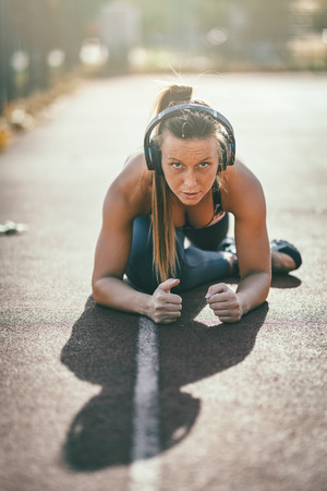 Young female runner, with headphones on her ears, doing stretching exercise on a public place, preparing for morning workout.の写真素材