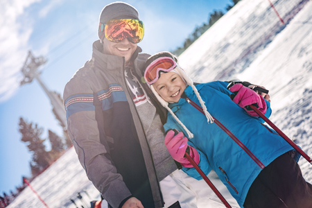 Beautiful young father and daughter enjoying in ski vacations. They are posing on sunny winter day and looking on camera with smile.の写真素材
