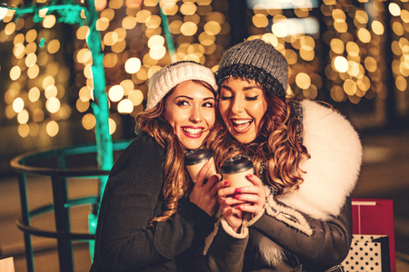 Two female friends enjoy the night out hugging and laughing with cups of coffee in their hands.の写真素材