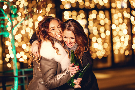 Two female friends enjoy the night out hugging and laughing with bottles of beer in their hands.の写真素材