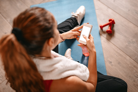 Attractive woman surfing on social media on a smartphone while taking break of exercising in living room at home.の写真素材