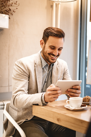 Handsome young smiling man drinking coffee in a cafe. He is surfing the internet on a digital tablet.の写真素材