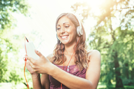 Woman relaxes with headphones listening to music sitting in park. She is doing selfie on her smartphoneの写真素材