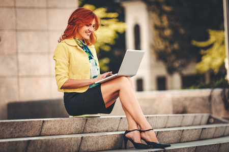 Smiling business woman is sitting on stairs in office district and working at the laptop.の写真素材