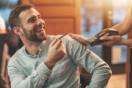 A handsome young man pays to waitress for coffee with credit card in a cafe.