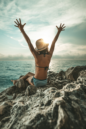 Beautiful little girl is sitting on the sea rock in the sunset. Her arms are high open and sunlight is shining through her hands.の写真素材