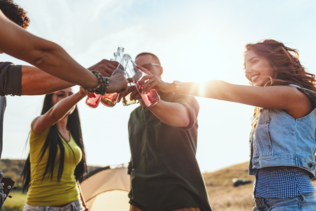 Happy young friends enjoy a sunny day in nature. They're clinking beer bottles, laughing and talking happy to be together.の写真素材