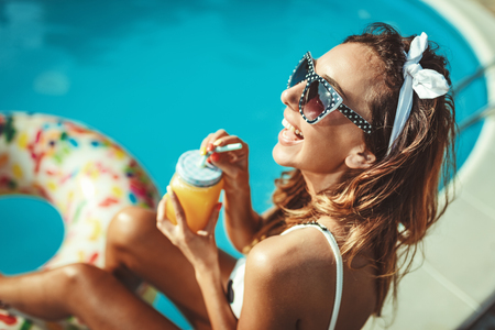 Beautiful smiling happy young woman enjoying sunbathing by swimming pool. She is sitting by clear blue water with a glass of cocktail.の写真素材