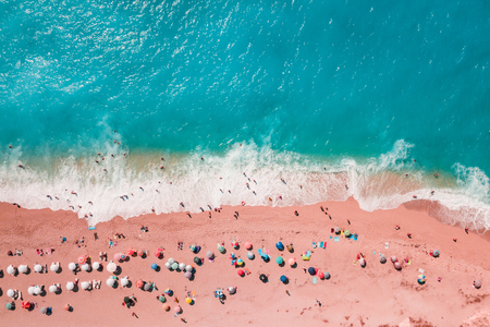 Soft blue waves of the sea with white foam on tropical sandy beach toned trendy living coral color of year 2019 with umbrellas shoot from drone.の写真素材