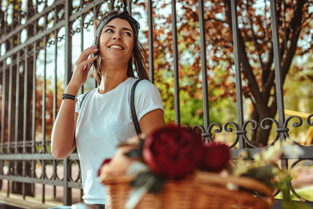 Laughing young woman talking on smartphone sitting on a fence in a sunny day, beside the bike with flower basket.の写真素材