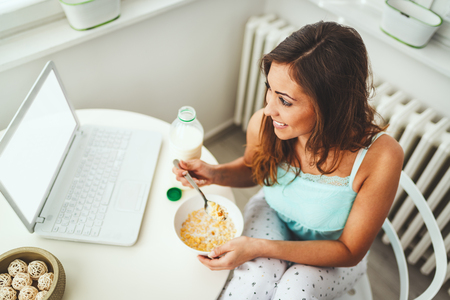 Beautiful young smiling woman is enjoying and relaxing in the morning, eating healthy breakfast and looking at laptop.の写真素材