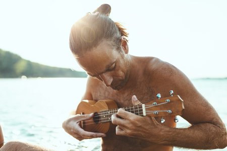 Happy young musician has a great time at the beach. He is sitting in the water, playing ukulele and singing. Sunset over water.の写真素材